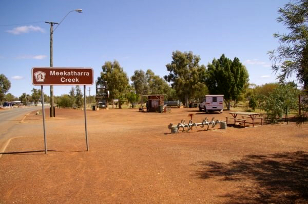 The road leading in to Meekathara. The earth is red, with tall trees, a bench and a caravan in the distance. The sign in the left foreground reads ‘Meekatharra Creek’. 